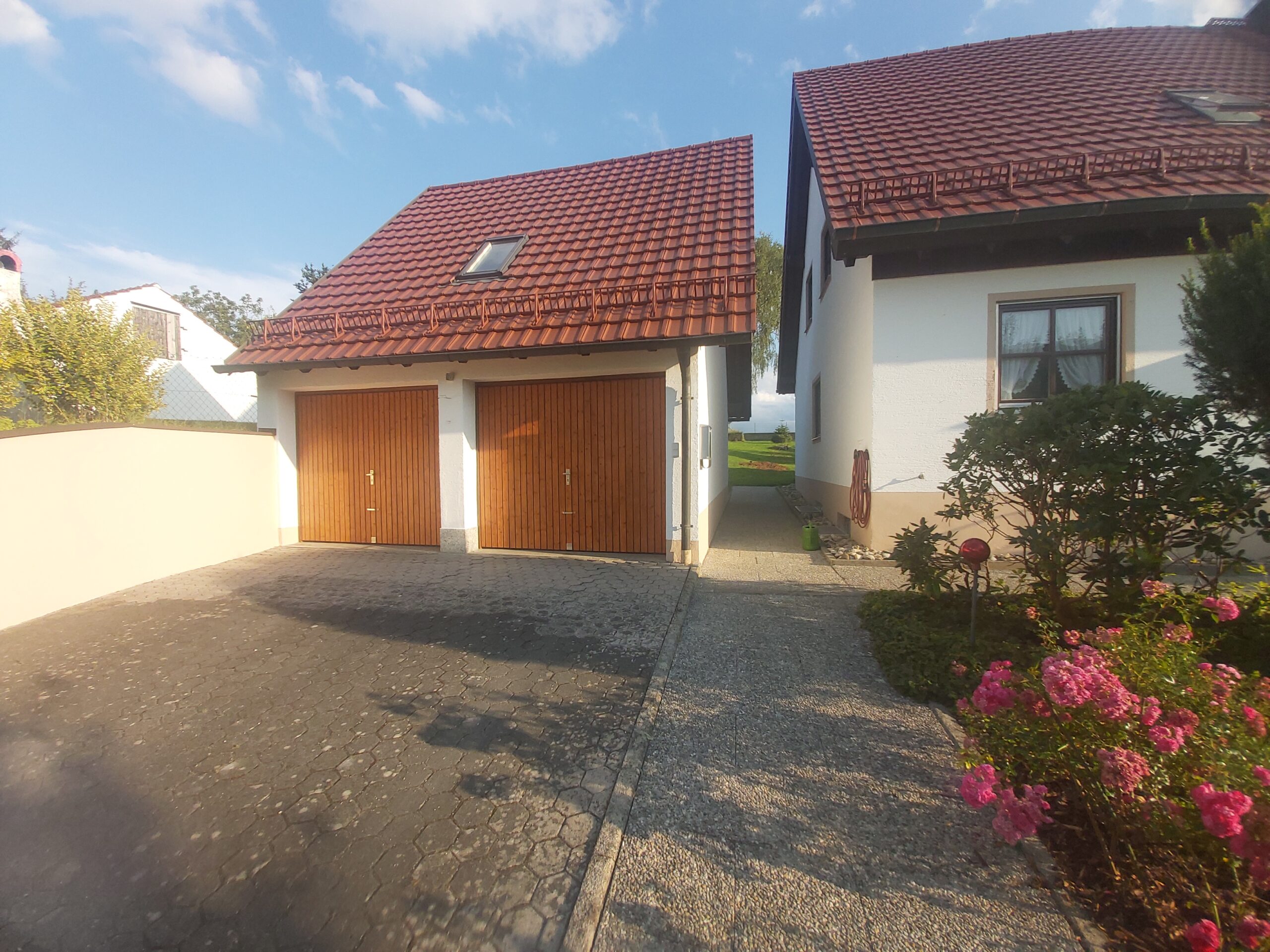 Widok na podjazd z dwustanowiskowym garażem o drewnianych bramach oraz sąsiedni dom jednorodzinny z ogrodem i kwiatam