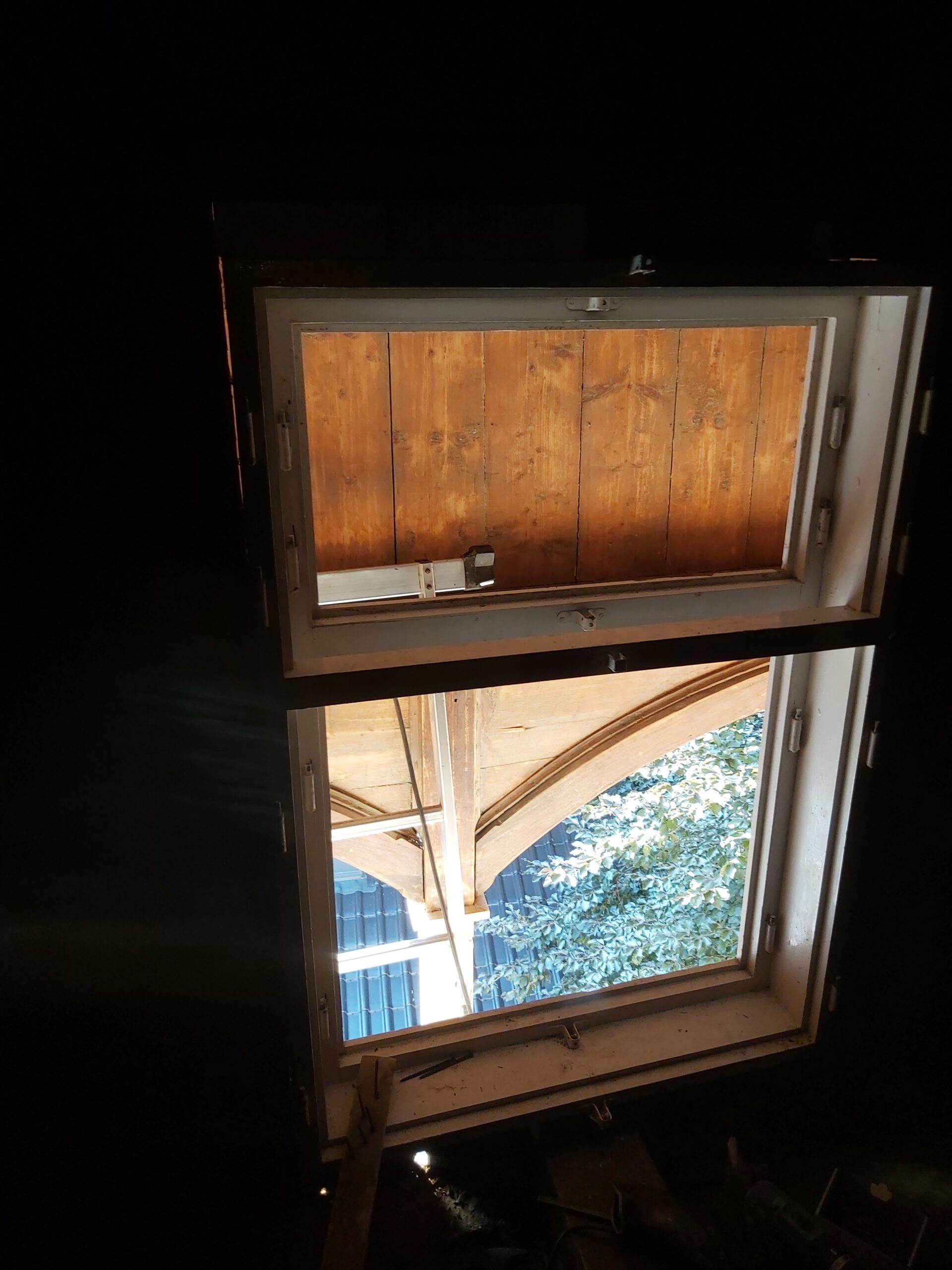 Widok od wewnątrz na dwuskrzydłowe okno w zabudowanym balkonie, z górnym skrzydłem zasłoniętym drewnianą okładziną i dolnym otwartym na zewnątrz podczas prac wykończeniowych.