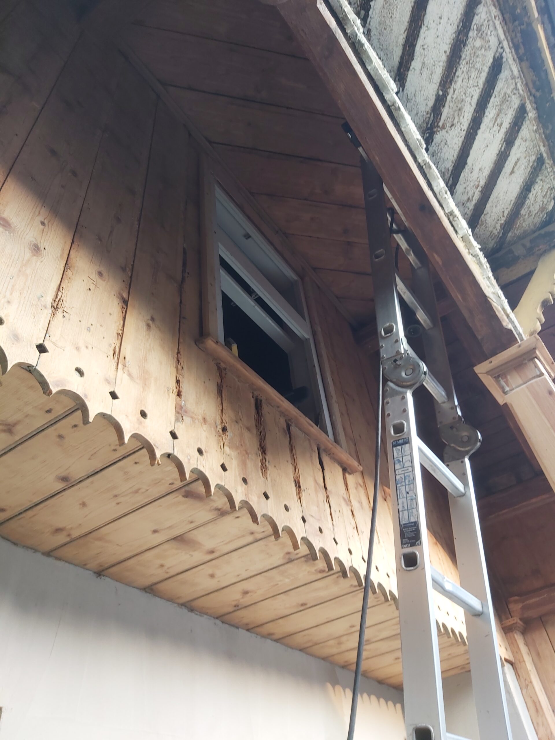 Drewniana elewacja zabudowanego balkonu z małym uchylonym oknem, dekoracyjną frezowaną podbitką oraz aluminiową drabiną ustawioną przy ścianie podczas prac wykończeniowych.