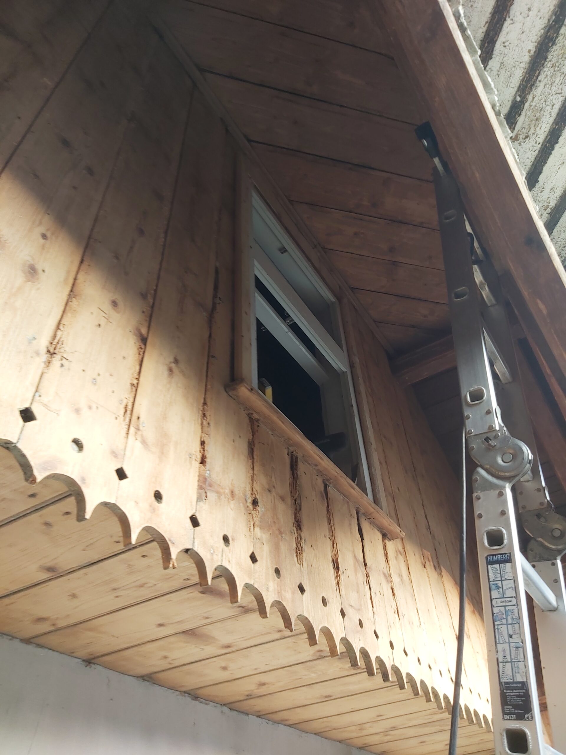 Drewniana elewacja zabudowanego balkonu z małym oknem uchylnym, dekoracyjną frezowaną podbitką oraz rozstawioną drabiną podczas prac wykończeniowych.