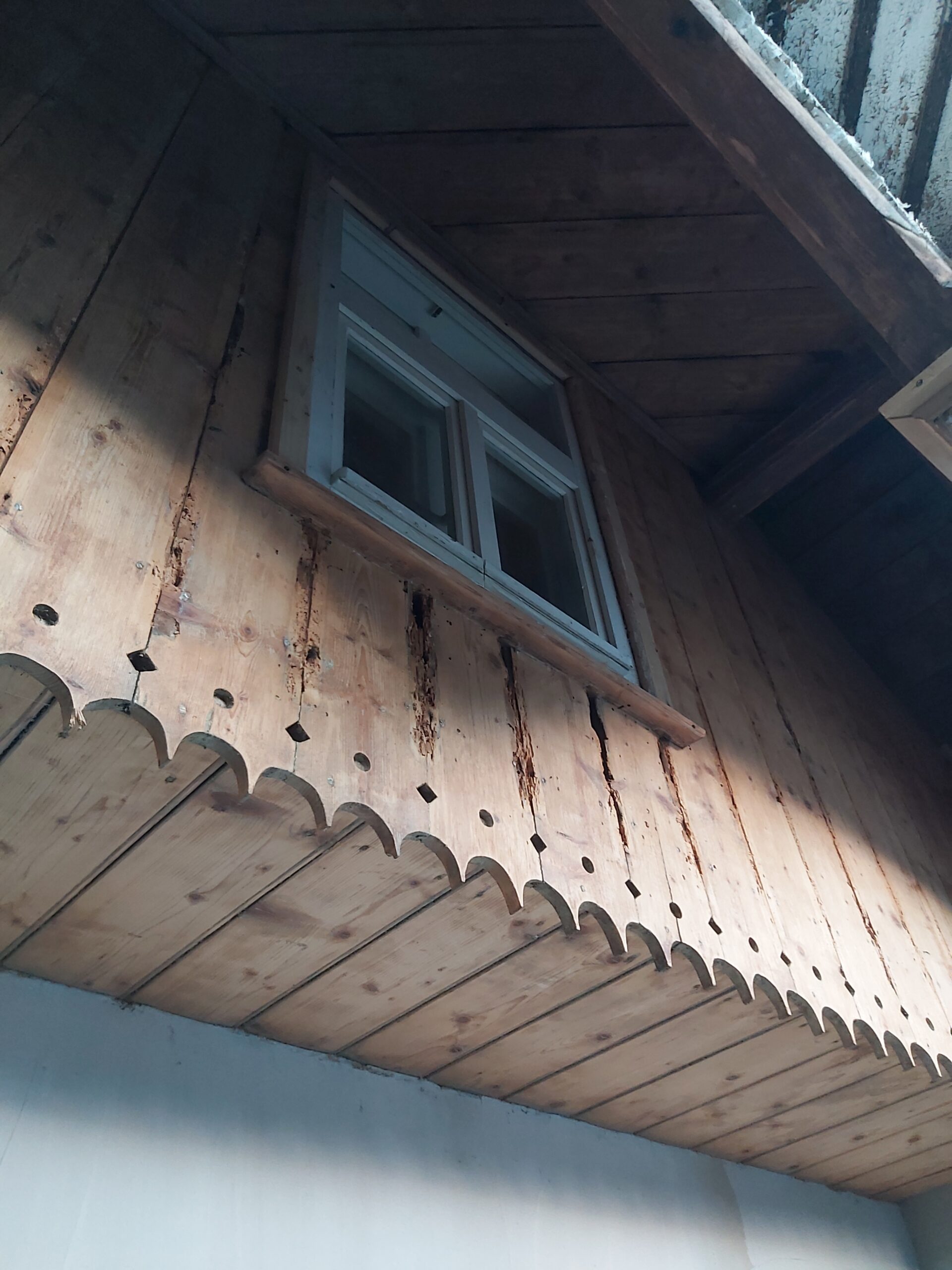 Drewniana elewacja zabudowanego balkonu z białym oknem, dekoracyjnym frezowanym podbitkiem oraz widocznymi śladami naturalnego starzenia drewna.