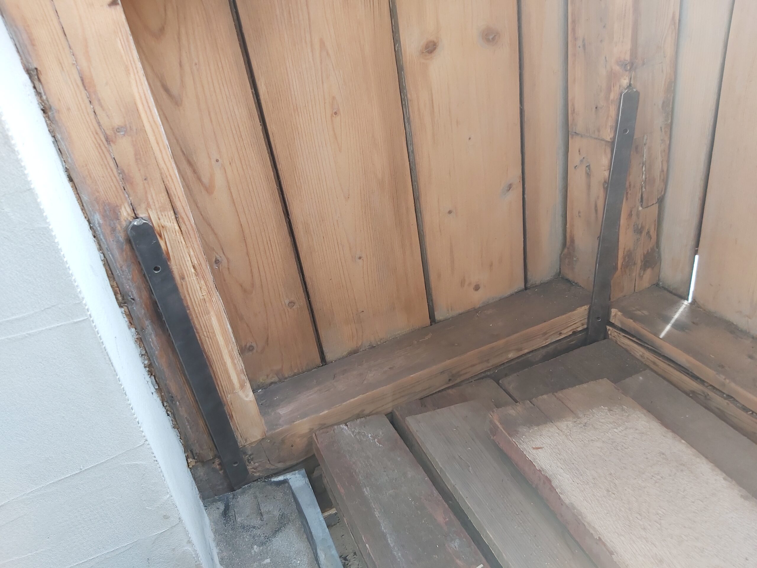 Detal narożnika drewnianej zabudowy balkonu – pionowe deski ścienne, metalowe kątowniki wzmacniające konstrukcję oraz fragment drewnianej podłogi w trakcie montażu.