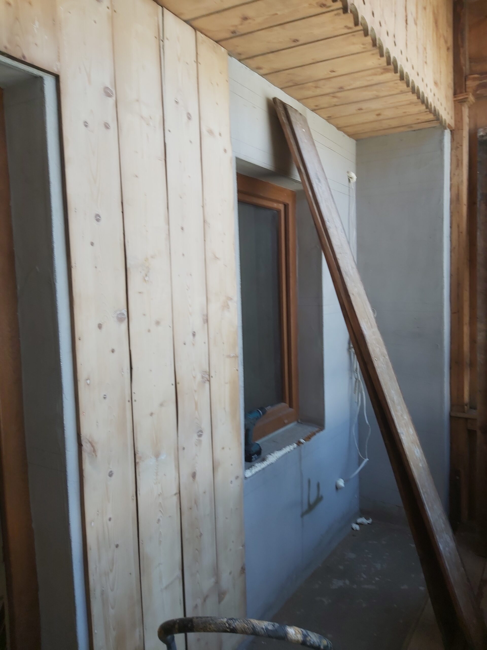 Montaż drewnianej zabudowy balkonu – jasne pionowe deski na ścianie, okno w drewnianej ramie oraz przygotowana powierzchnia pod dalsze prace wykończeniowe.