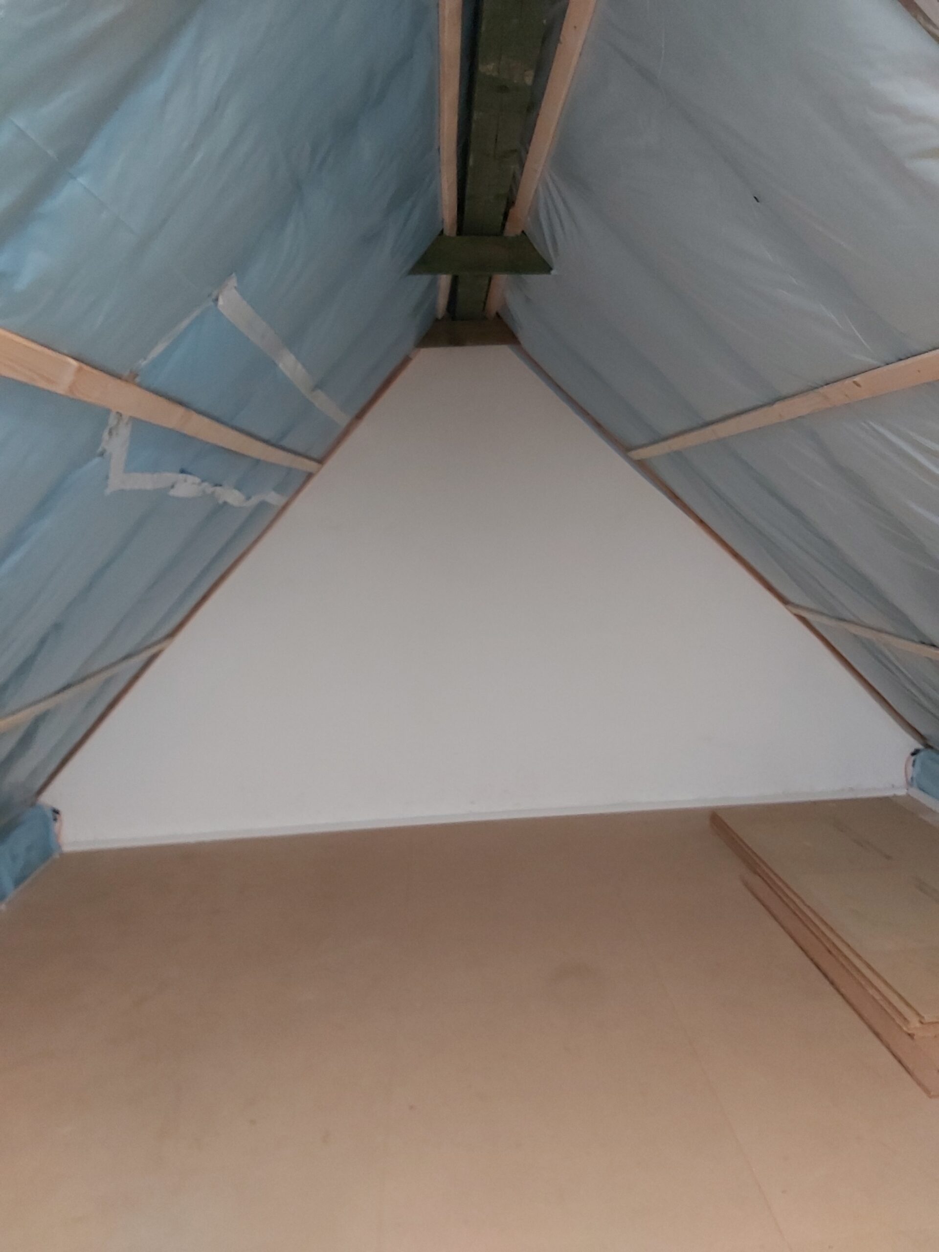 Widok na ścianę szczytową poddasza po zabudowie, z widoczną folią paroizolacyjną pod połaciami dachowymi oraz przygotowaną podłogą z płyt.