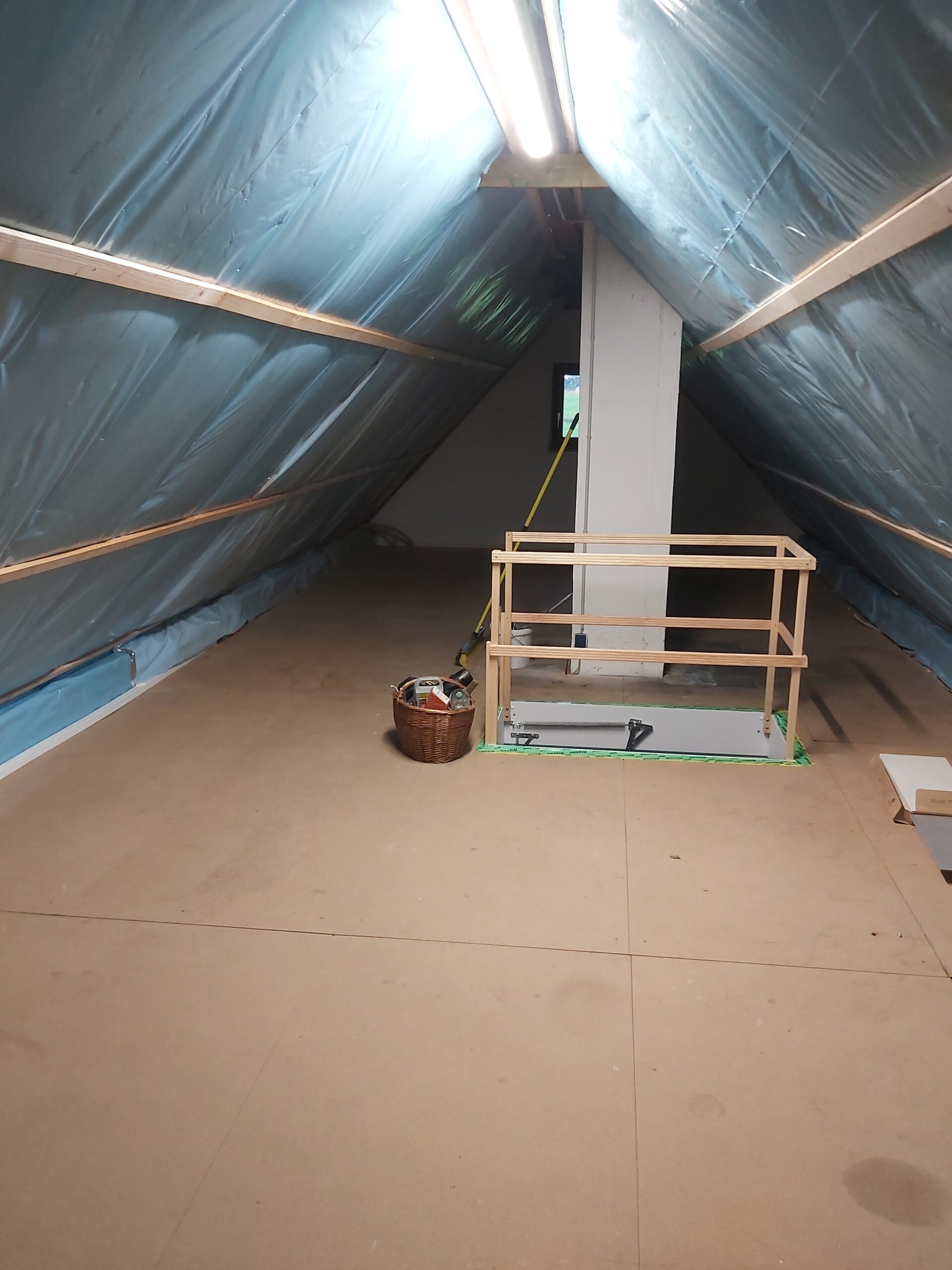 Widok na poddasze w trakcie prac remontowych, z widoczną folią izolacyjną pod połaciami dachowymi, surową podłogą z płyt oraz tymczasowym zabezpieczeniem otworu schodowego.