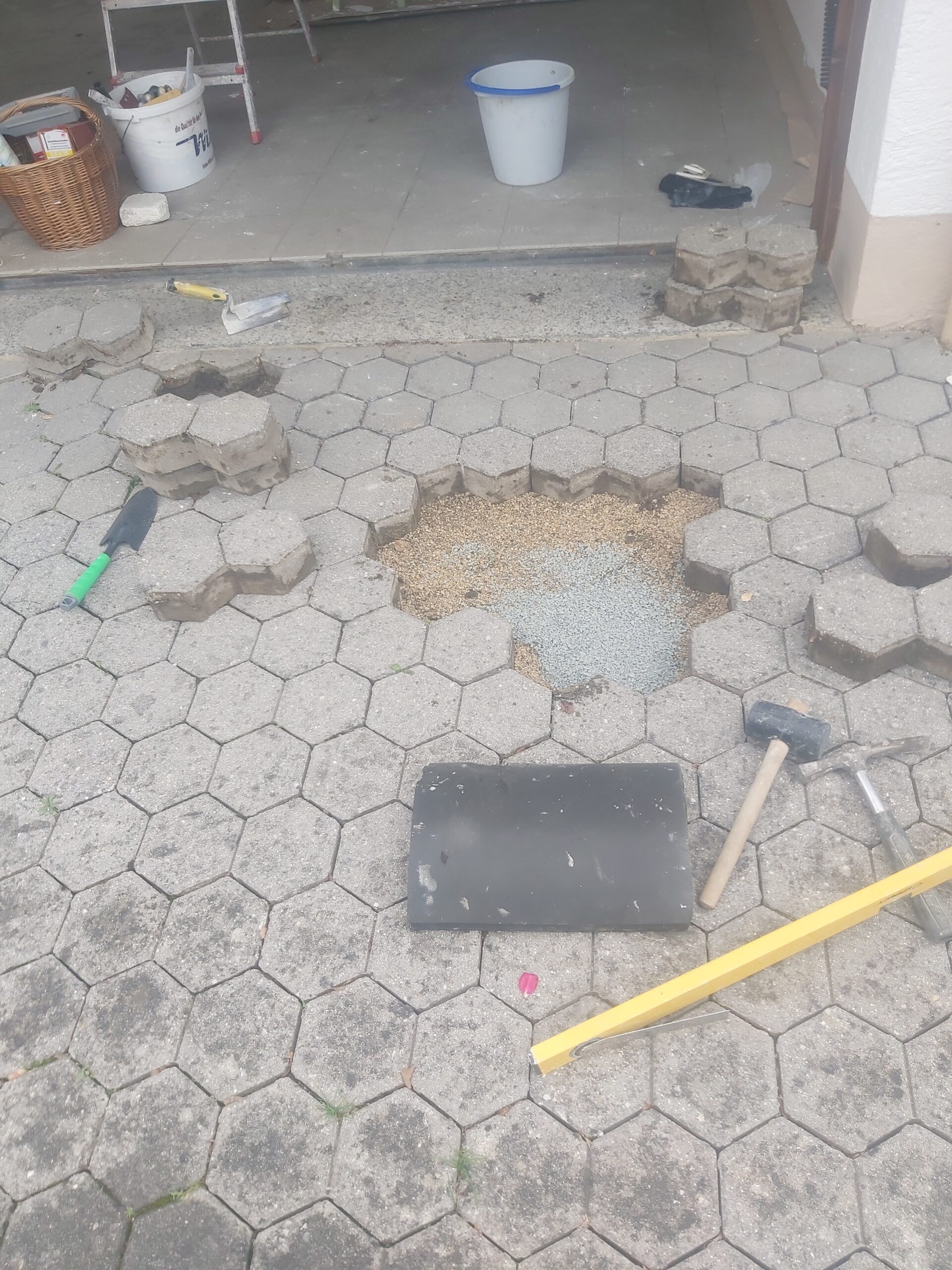 Otwarty fragment nawierzchni z kostki brukowej przed garażem, z odsłoniętym podłożem i narzędziami używanymi podczas prac naprawczych.