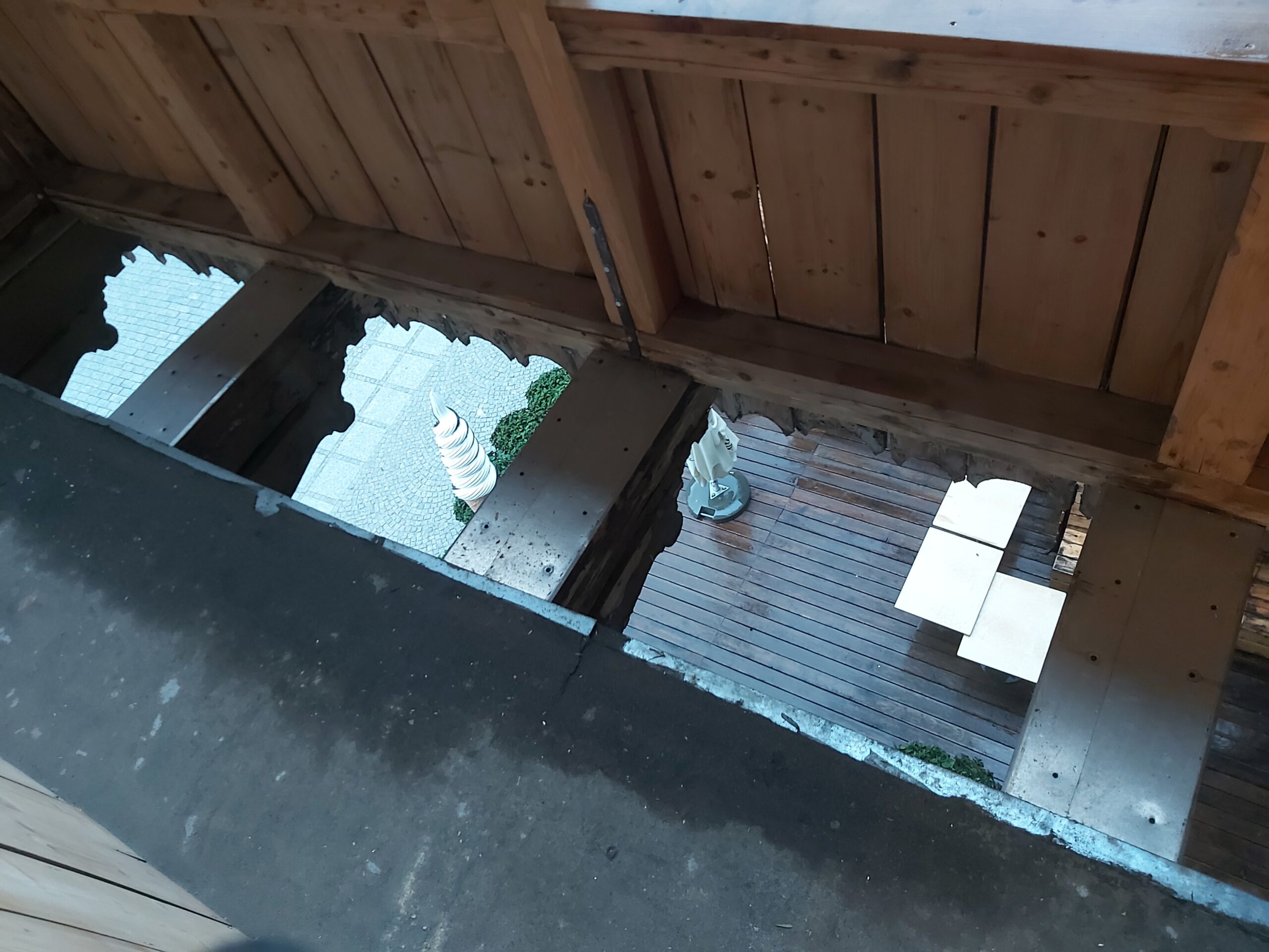 Otwarta podłoga balkonu w trakcie remontu, widoczne otwory po zdemontowanych deskach oraz odsłonięte belki konstrukcyjne z widokiem na taras poniżej.