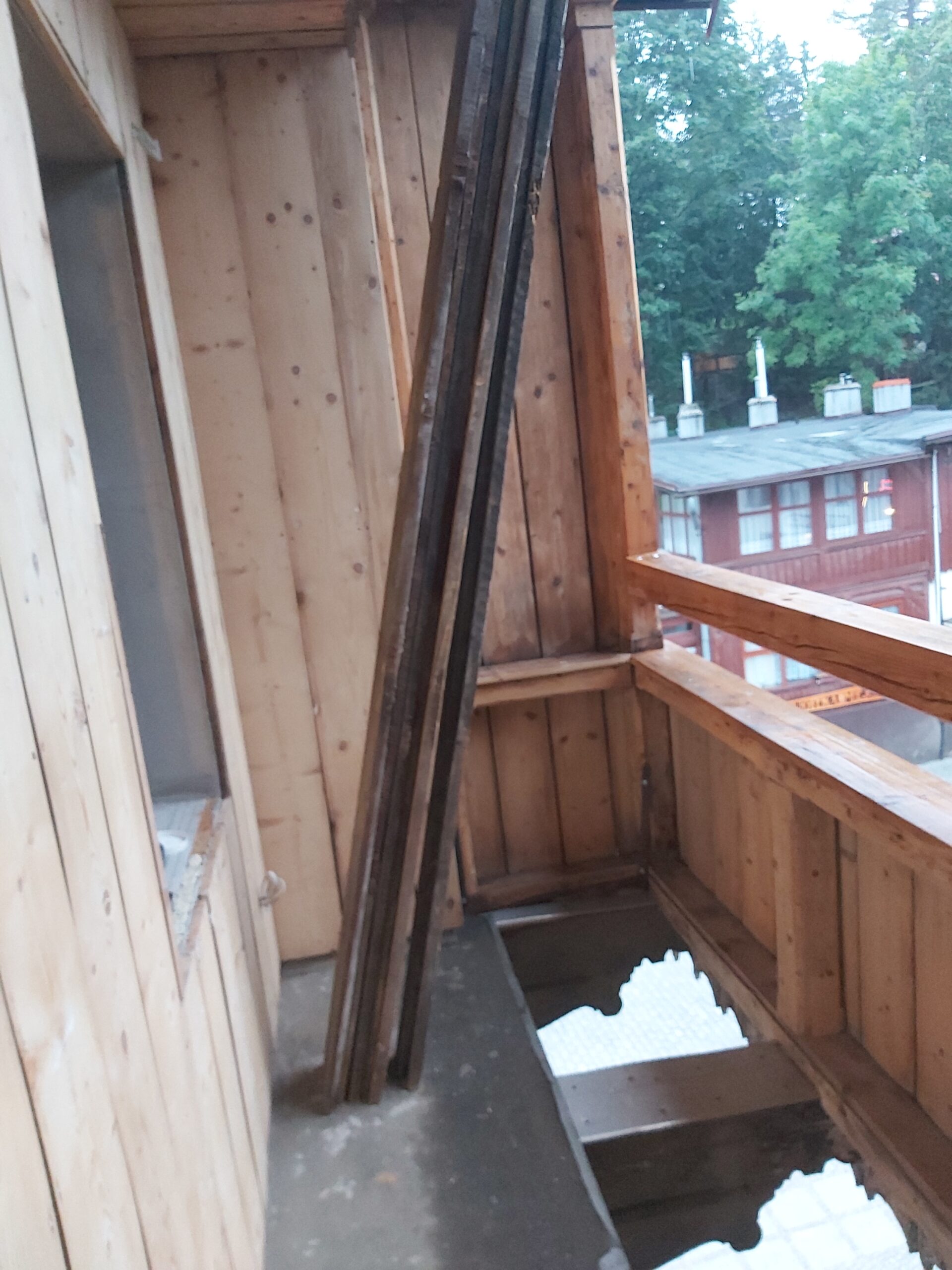 Balkon w trakcie remontu, widoczne zdjęte deski podłogowe oparte o ścianę oraz otwory w podłodze z odsłoniętą konstrukcją nośną.