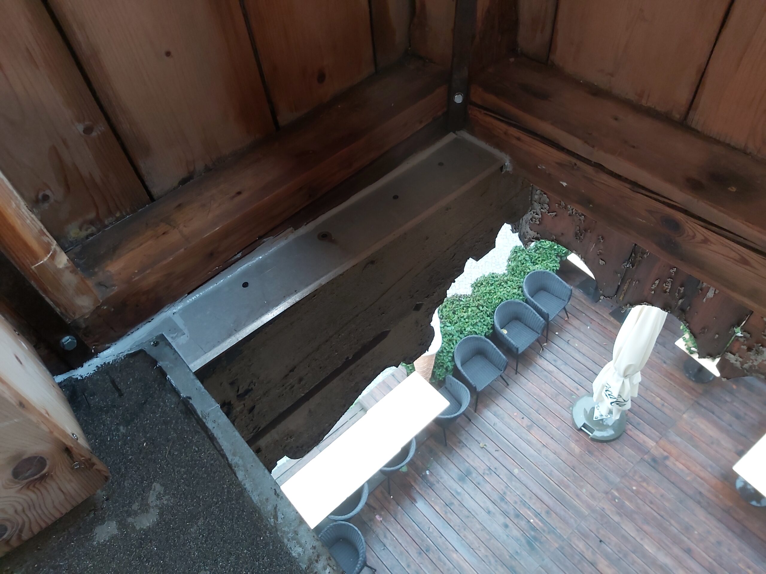 Otwór w podłodze balkonu przy narożniku, z widoczną drewnianą konstrukcją, metalowym wzmocnieniem oraz widokiem na taras poniżej.