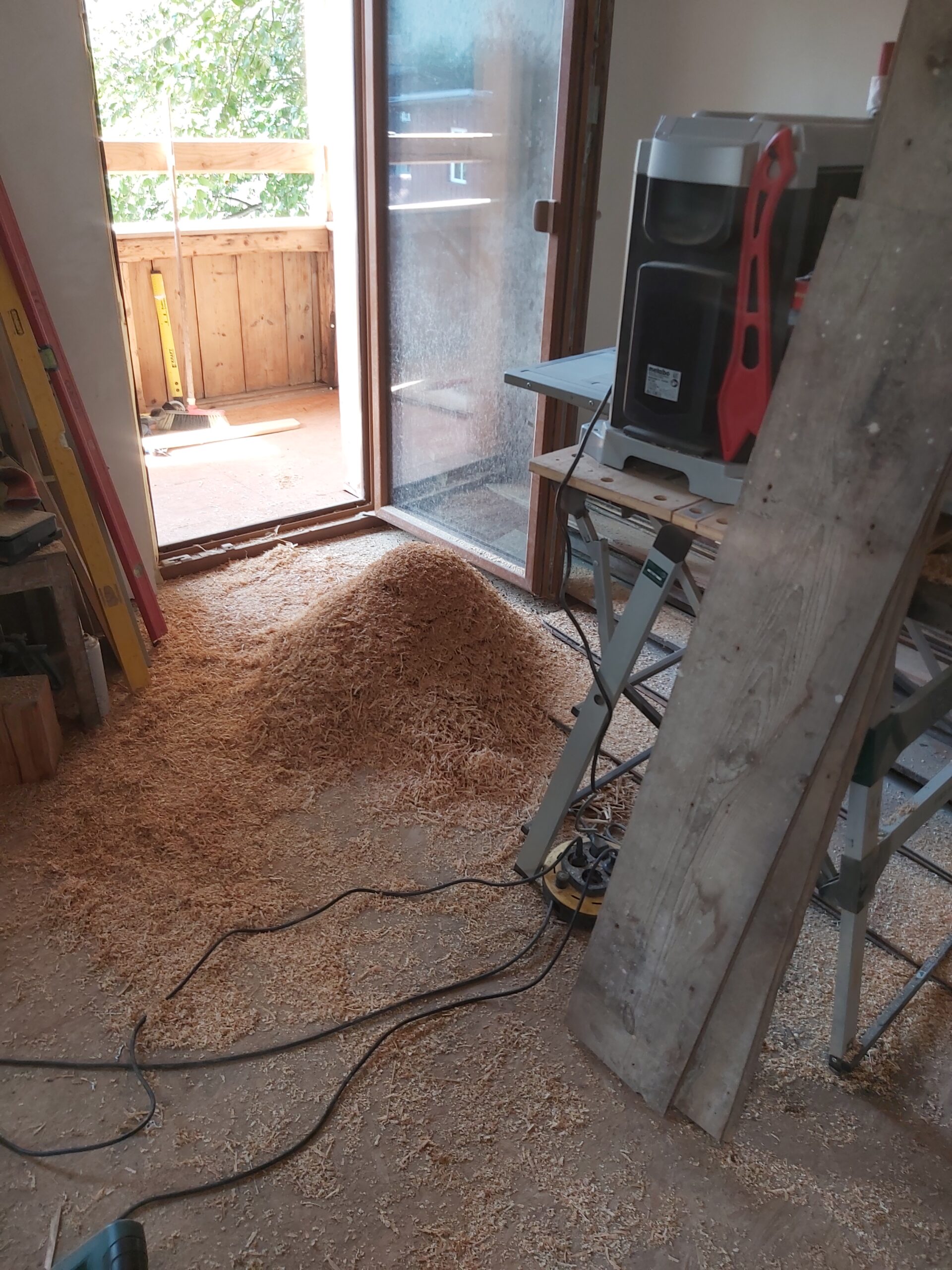 Wnętrze pomieszczenia podczas prac stolarskich, na podłodze leży sterta trocin, w tle otwarte drzwi balkonowe i maszyna do obróbki drewna.