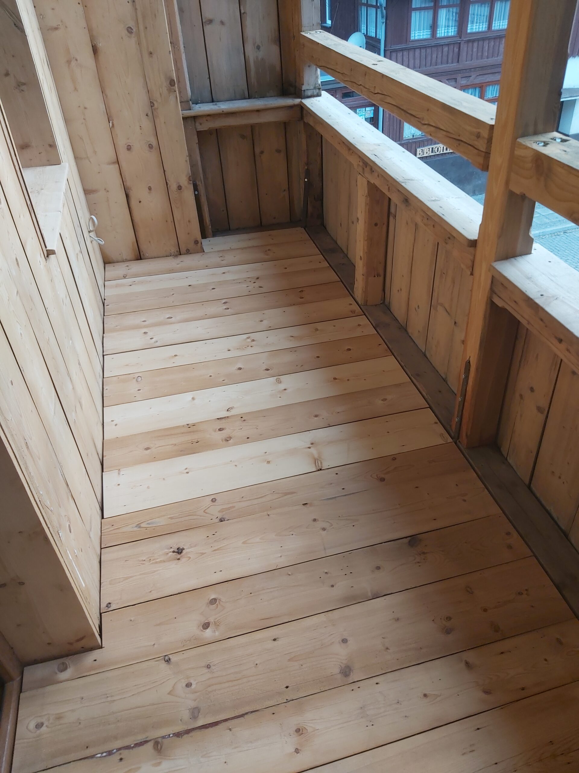 Drewniana podłoga na balkonie z jasnych desek, ułożonych równolegle, otoczona drewnianą balustradą i ścianami.