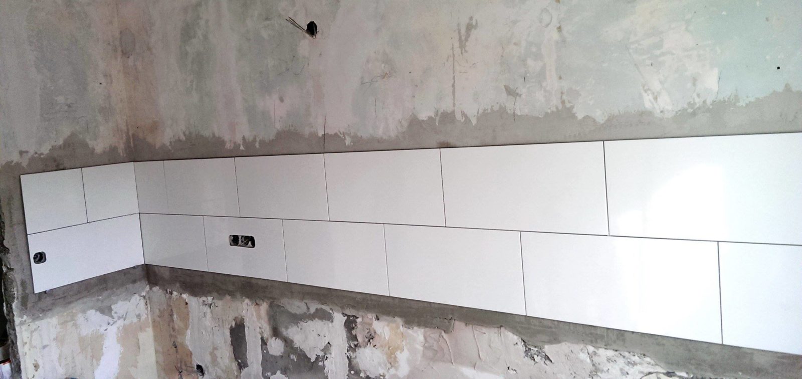 Białe płytki ścienne ułożone pasem w łazience, widoczna surowa ściana powyżej oraz przygotowane otwory instalacyjne.