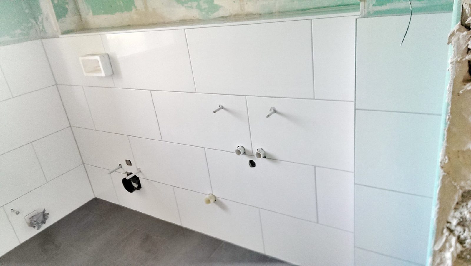 Biała ściana łazienki wyłożona płytkami, widoczne podejścia hydrauliczne i instalacyjne przygotowane pod montaż armatury i zabudowy.