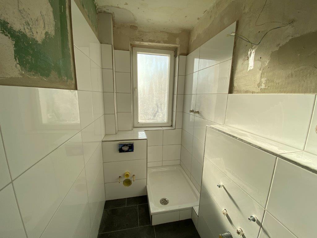 Mała łazienka w trakcie remontu, białe płytki ścienne, zamontowany brodzik prysznicowy i instalacje sanitarne pod armaturę.