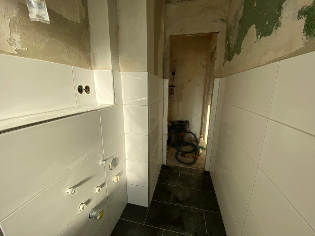 Wąska łazienka z białymi płytkami ściennymi, widoczne podejścia instalacyjne oraz wnęka prowadząca do pomieszczenia w trakcie remontu.