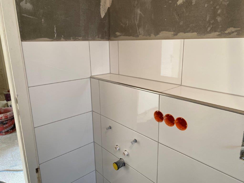 Biała ściana łazienki wyłożona płytkami, wnękowa półka z płytek oraz przygotowane otwory pod instalację armatury.