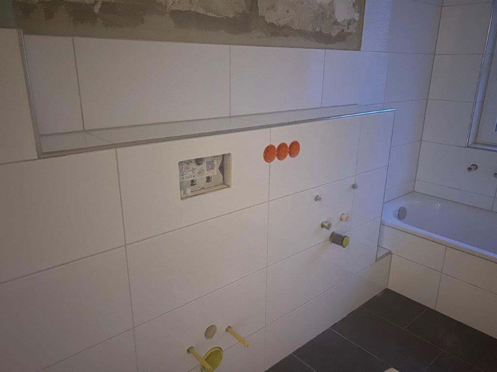 Ściana łazienki wykończona białymi płytkami, przygotowane otwory pod armaturę, spłuczkę podtynkową i wannę.