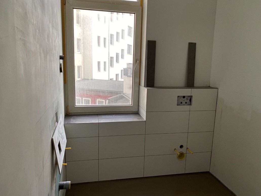 Ściana łazienki z oknem, białe płytki ceramiczne oraz przygotowane podejścia instalacyjne pod armaturę i zabudowę.