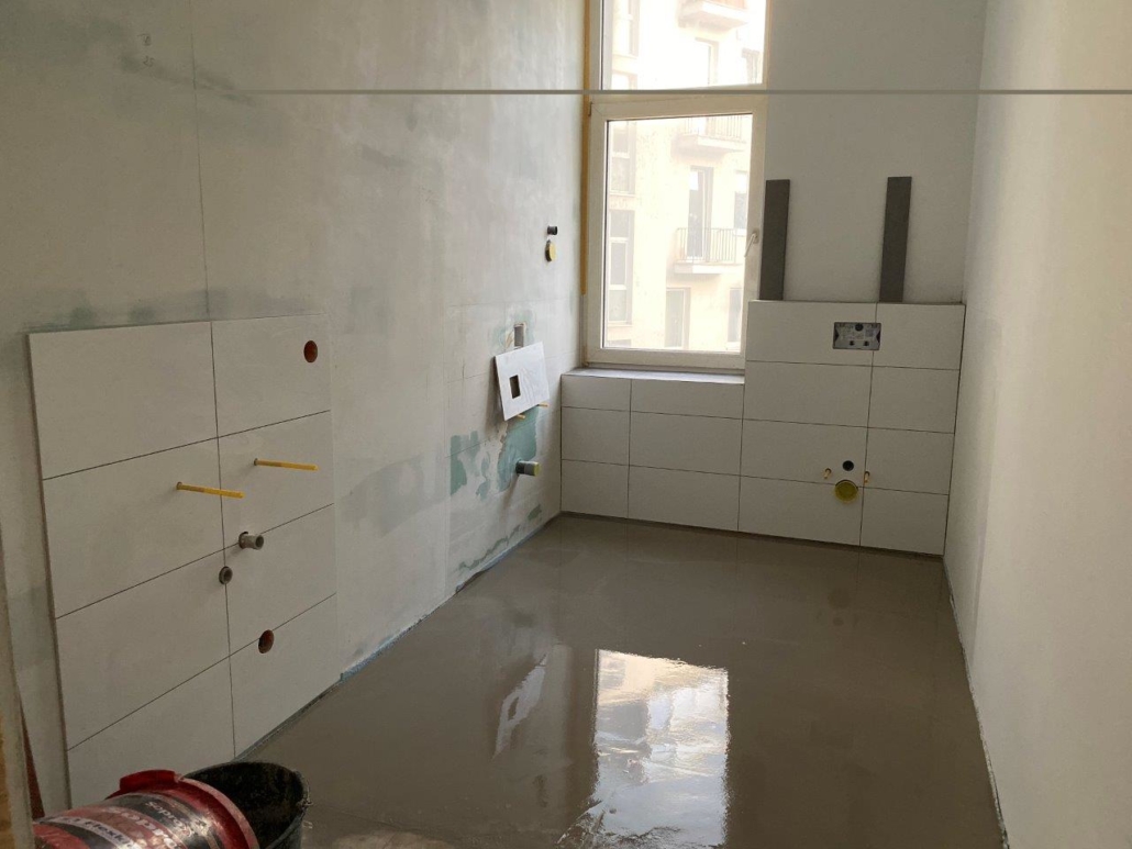 Świeża wylewka samopoziomująca w łazience, białe płytki ścienne z przygotowanymi wyjściami instalacyjnymi oraz okno doświetlające pomieszczenie.