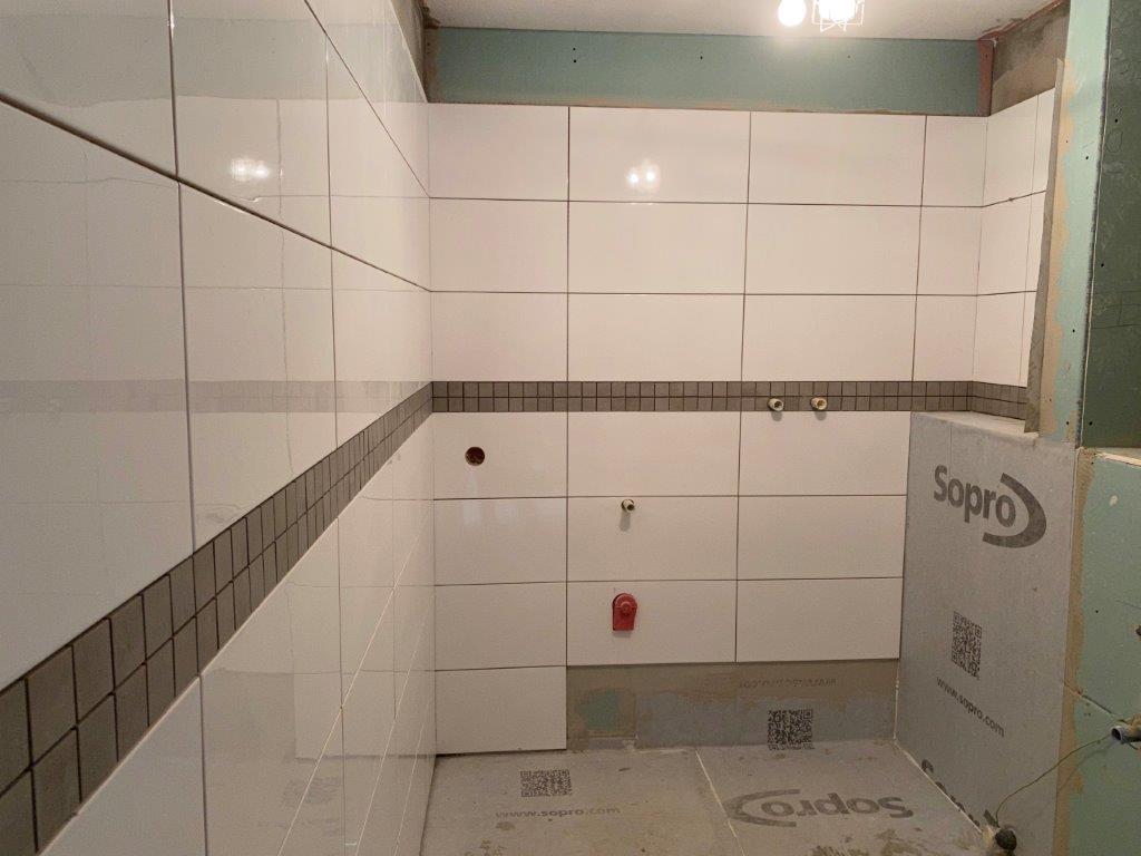 Białe płytki ścienne w łazience z poziomym pasem szarej mozaiki oraz widocznymi wyjściami instalacji.