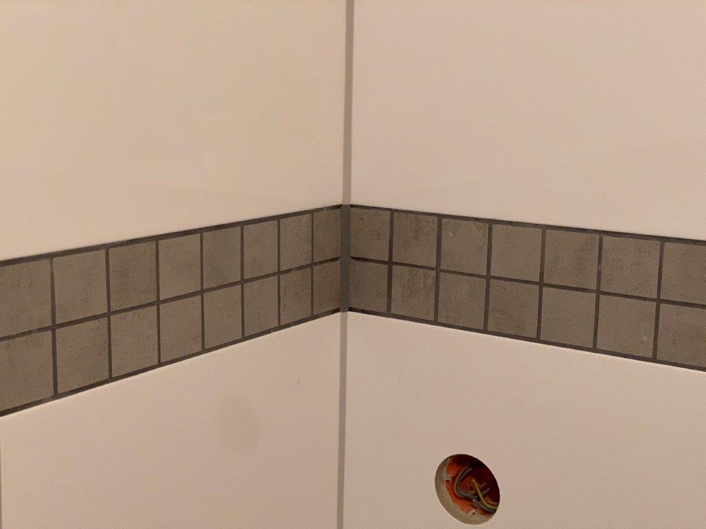 Narożnik ściany w łazience z białymi płytkami i szarym pasem dekoracyjnym z drobnej mozaiki.