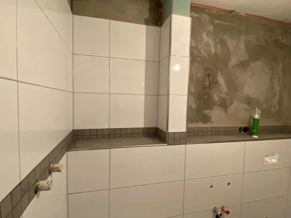 Wnęka prysznicowa z półką wyłożoną białymi płytkami, szarym pasem dekoracyjnym oraz widoczną surową ścianą nad glazurą.