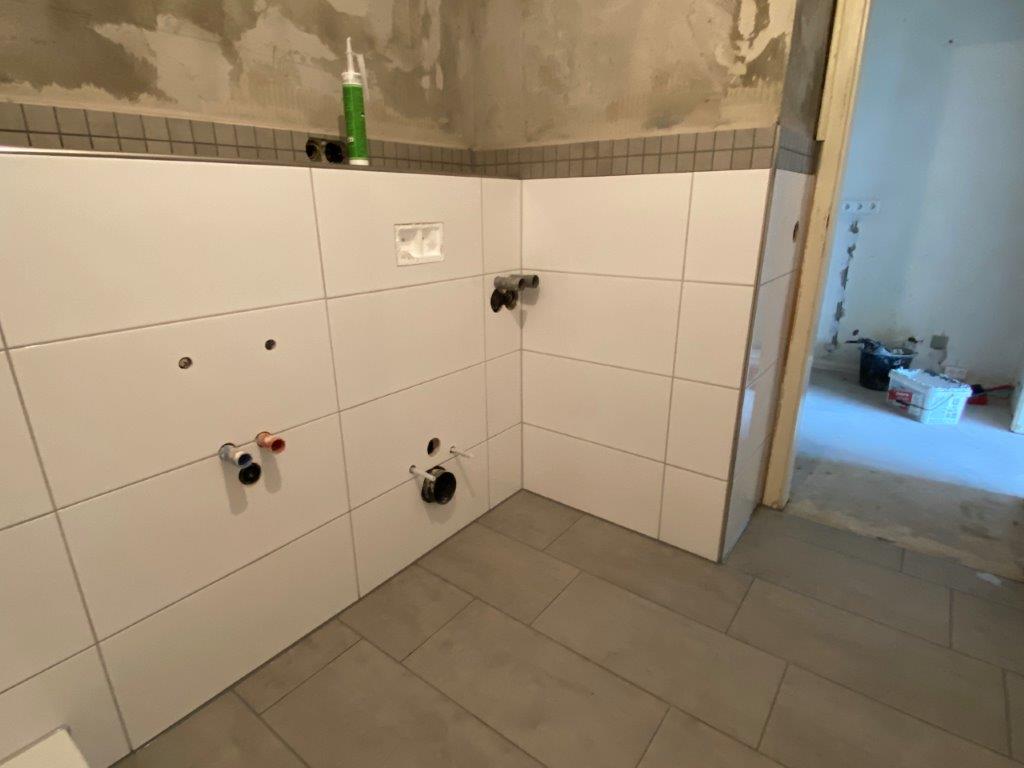 Ściana w łazience wyłożona białymi płytkami, widoczna instalacja podtynkowa prysznica oraz szara podłoga z płytek.