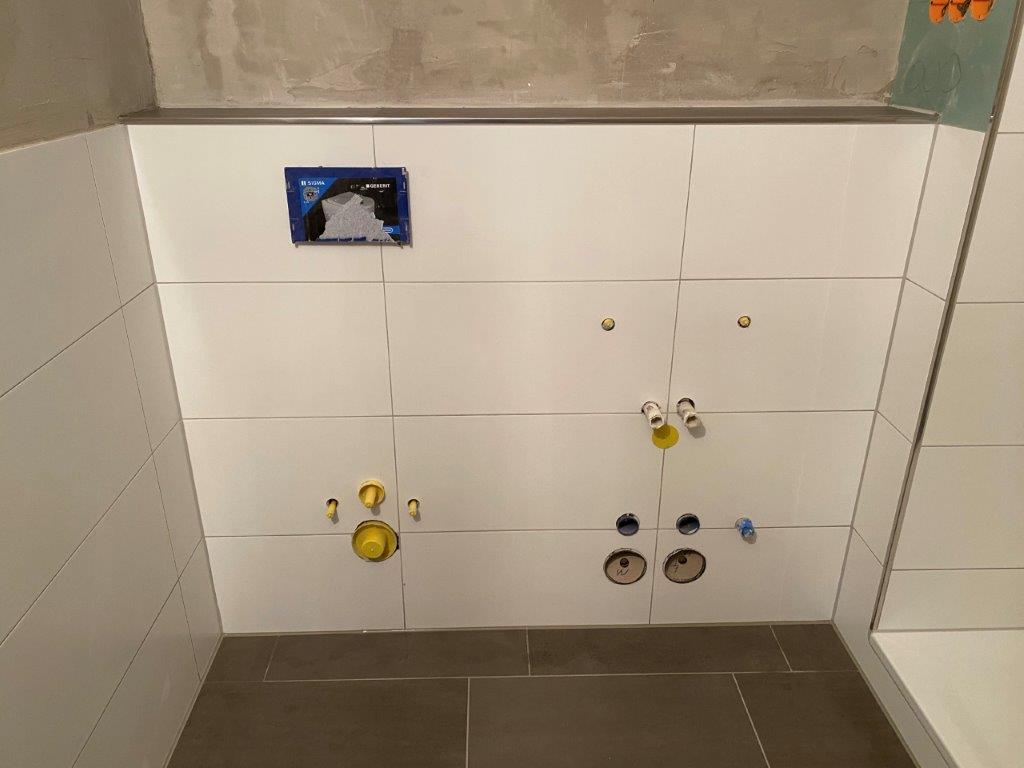 Ściana łazienki z zamontowanymi przyłączami hydraulicznymi i stelażem pod armaturę, wykończona białymi płytkami.