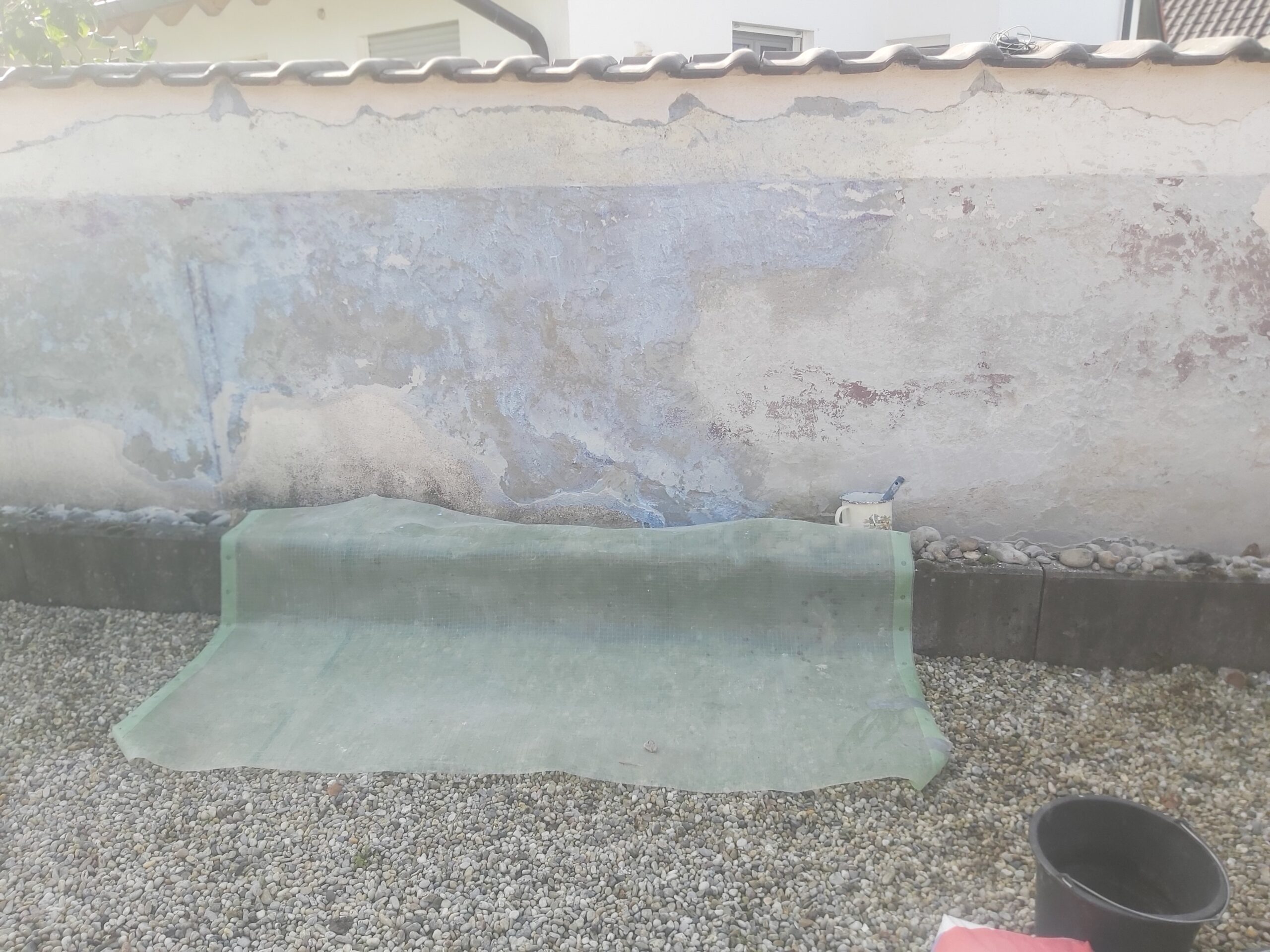 Rozłożona warstwa izolacyjna na żwirowym podłożu wzdłuż muru ogrodzeniowego, przygotowująca teren do dalszych prac wykończeniowych.