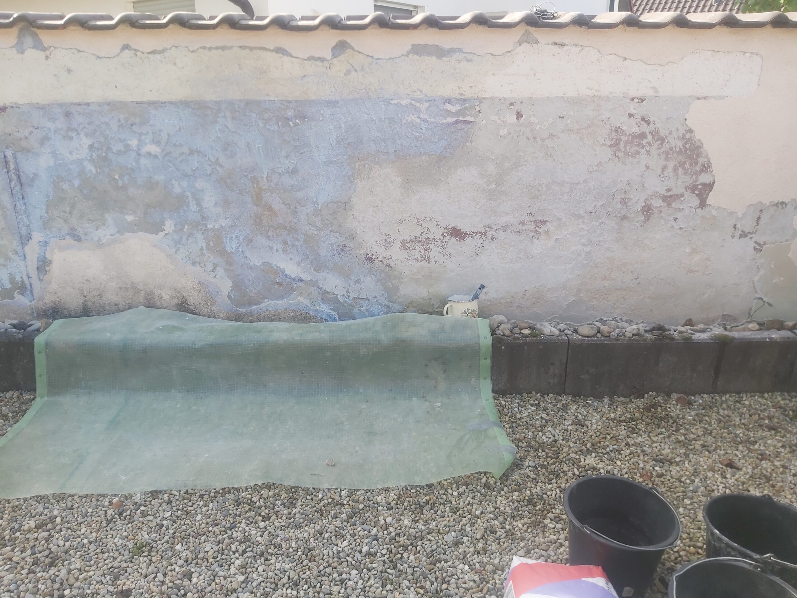 Fragment terenu przy murze ogrodzeniowym z rozłożoną warstwą izolacyjną na żwirowym podłożu, przygotowanym do dalszych prac wykończeniowych.