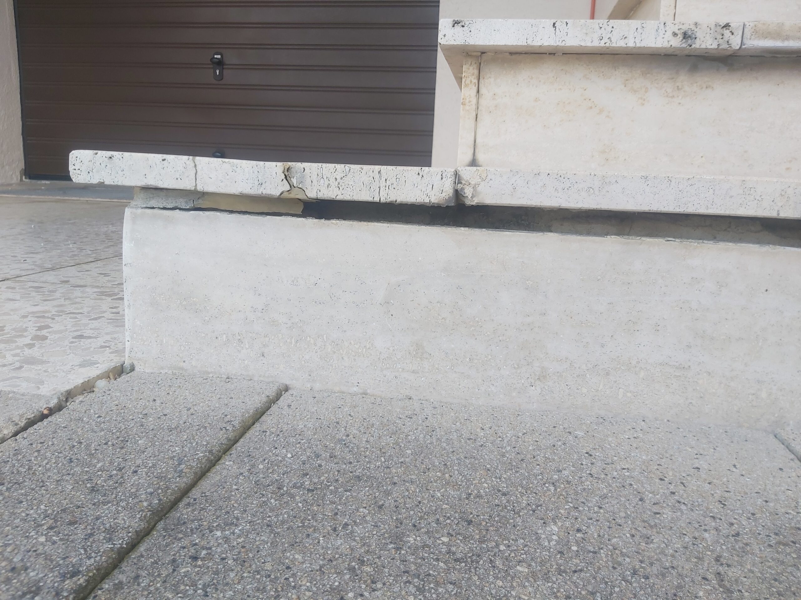 Widok na uszkodzony cokół przy wejściu do budynku, z widocznym pęknięciem i zabrudzeniami na styku betonu, kamienia oraz nawierzchni chodnika.