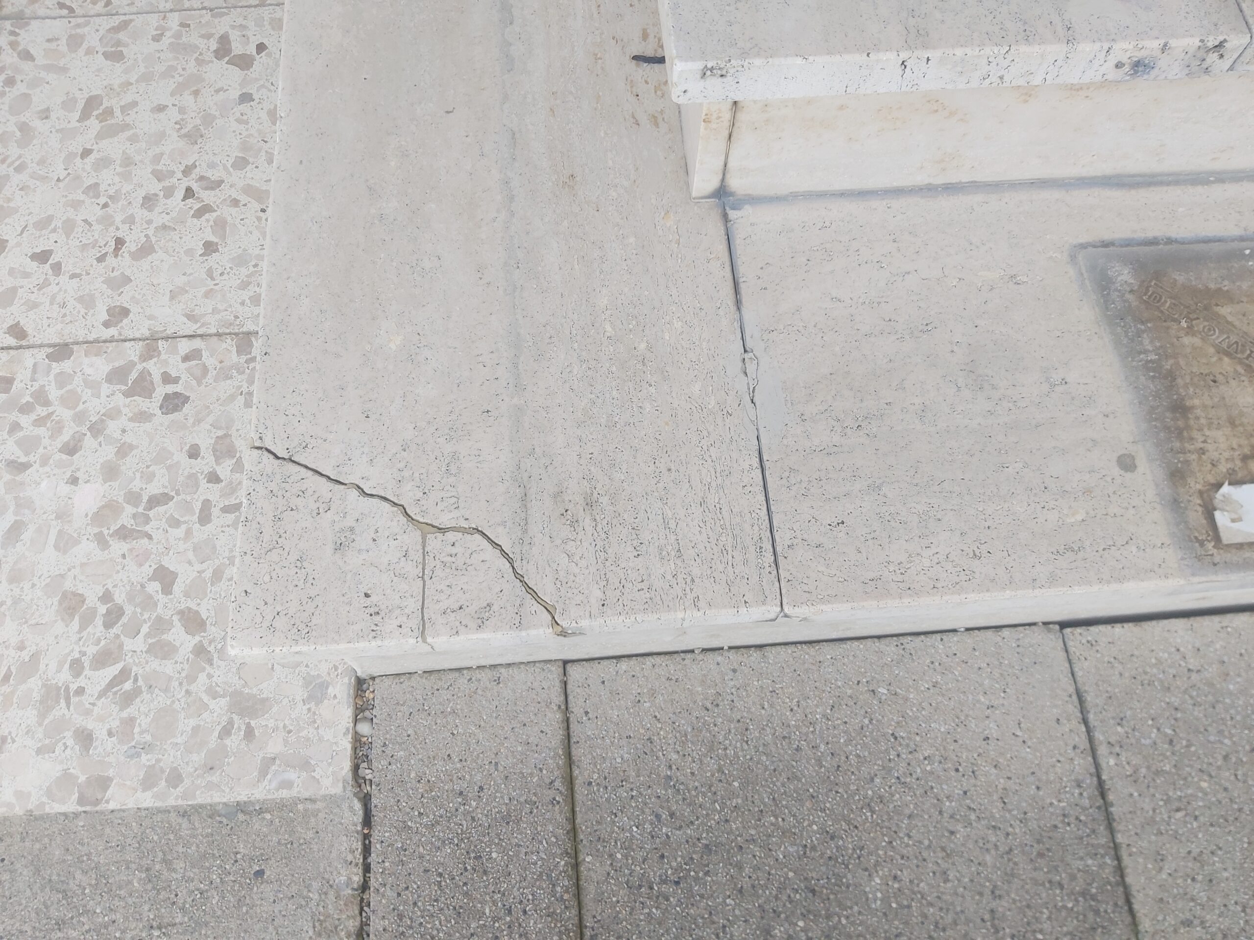 Zbliżenie na pękniętą płytę kamienną przy krawędzi schodów i chodnika, widoczne uszkodzenie powierzchni oraz naturalne ślady użytkowania.