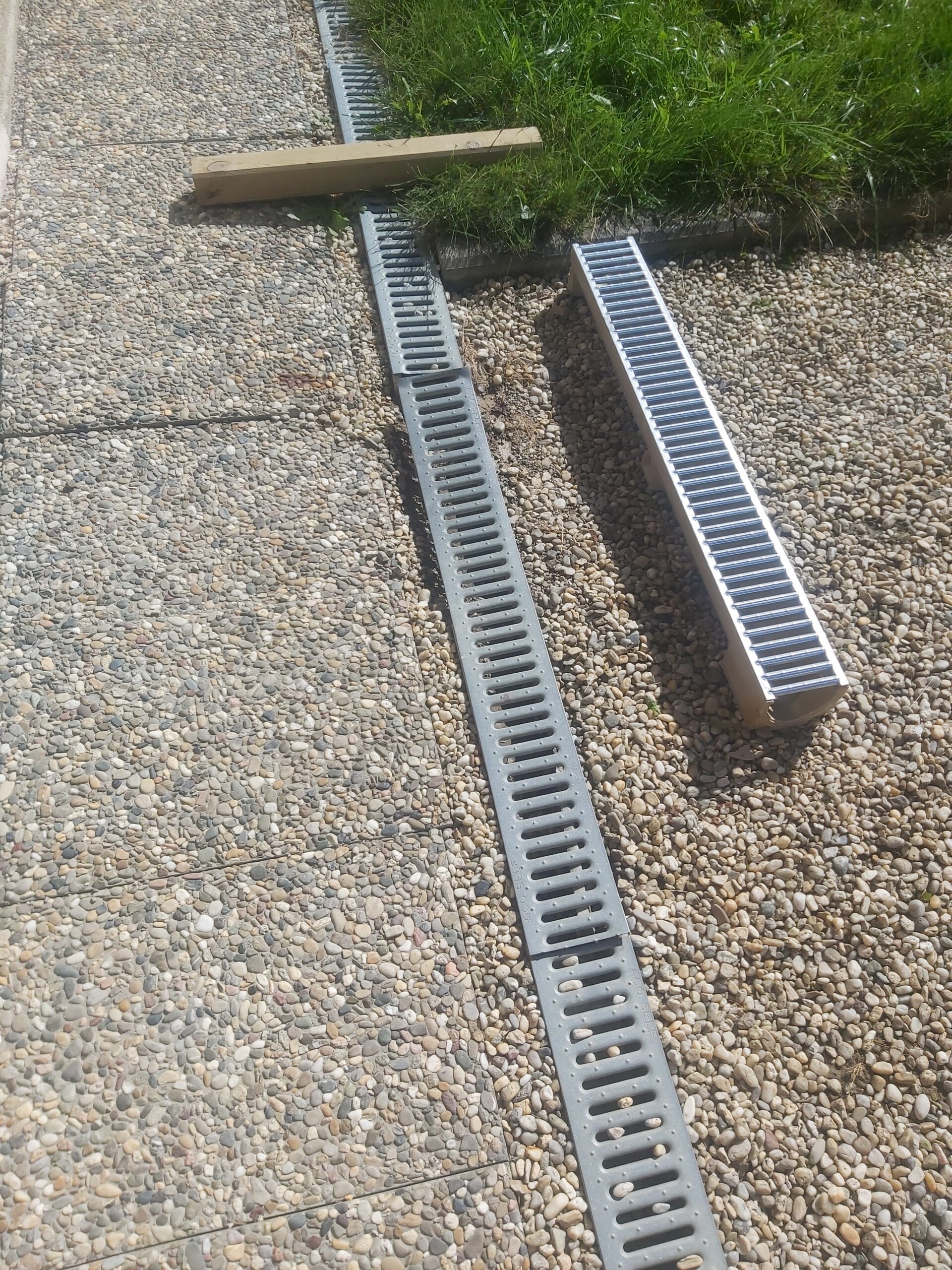 Elementy systemu odwodnienia liniowego ułożone na żwirowej nawierzchni, pokazujące przygotowanie do montażu przy ciągu komunikacyjnym.