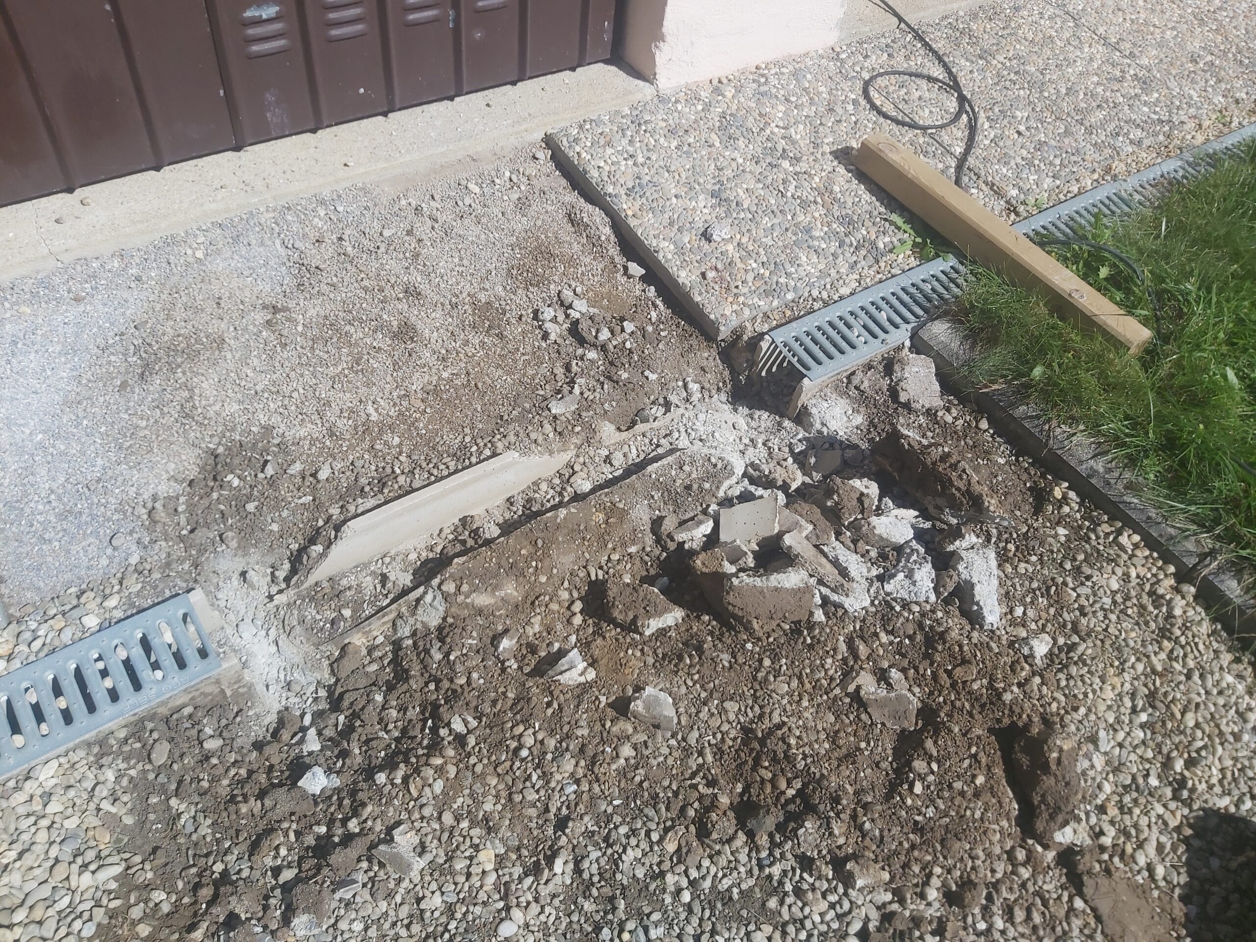 Fragment wjazdu do garażu w trakcie demontażu odwodnienia liniowego, z odsłoniętym gruntem, usuniętymi elementami betonu i przygotowaniem do ponownego montażu.