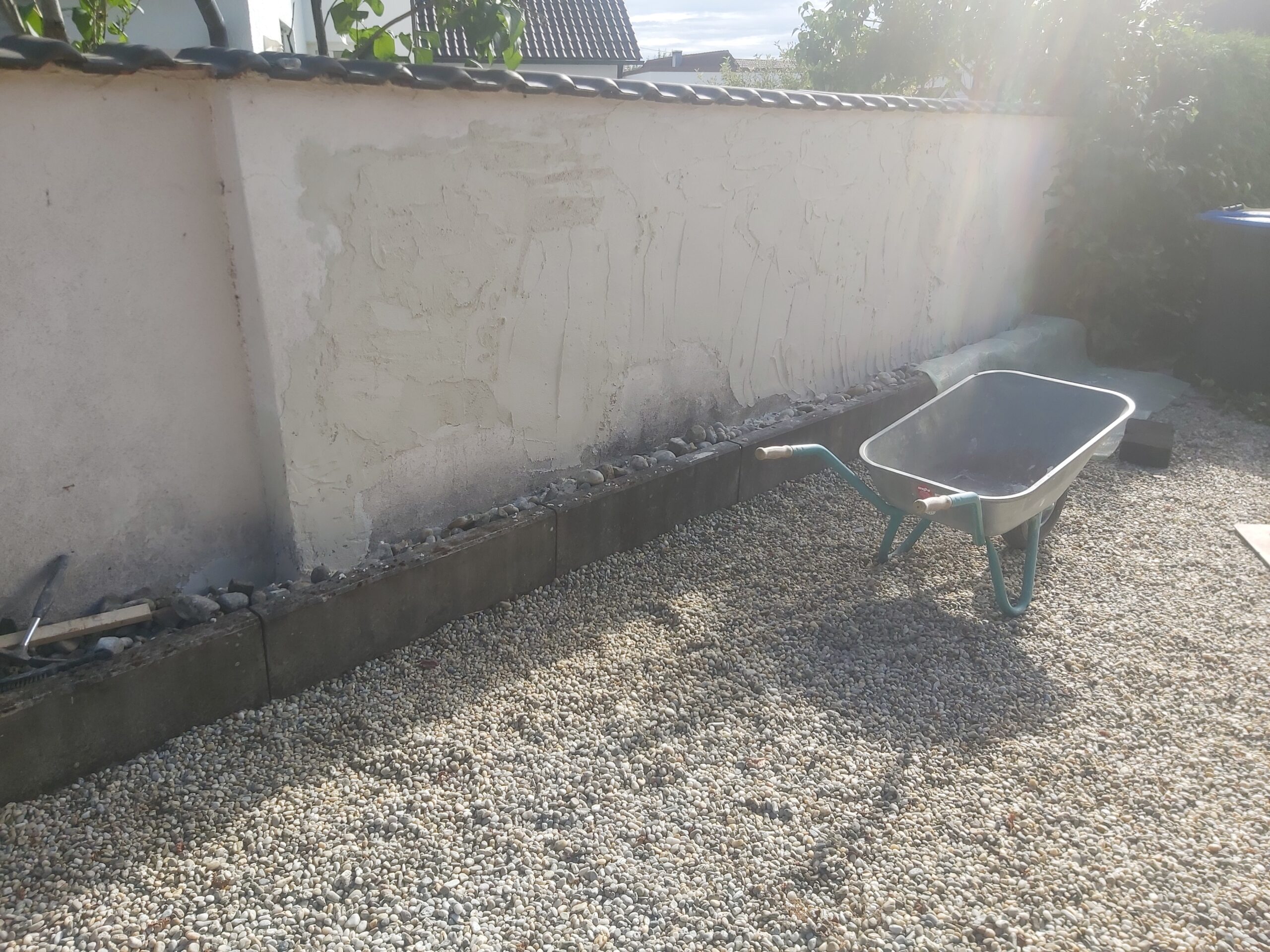 Żwirowa nawierzchnia wzdłuż muru ogrodzeniowego w trakcie prac porządkowych, z widoczną taczką i wyrównanym terenem wokół posesji.