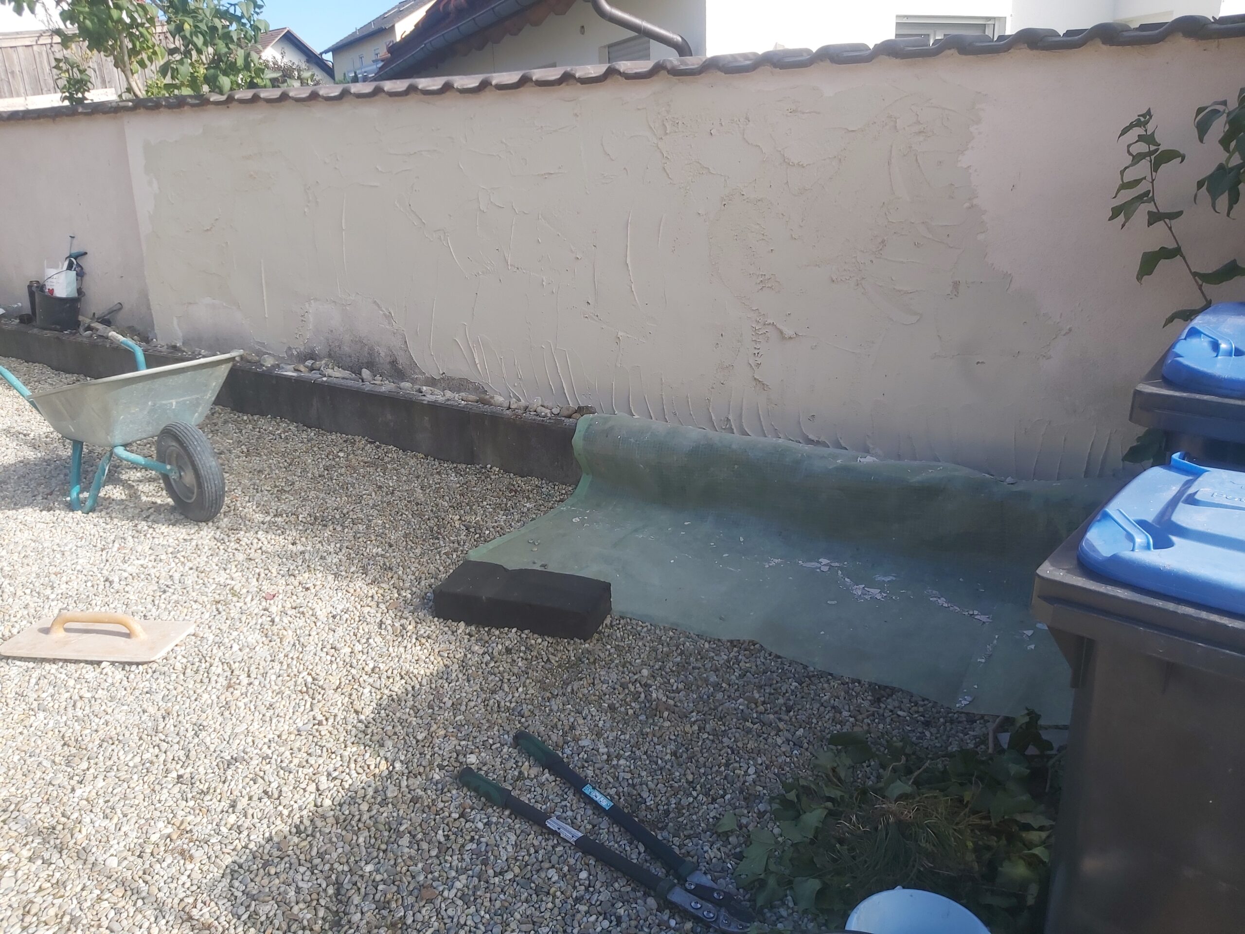 Teren przy murze ogrodzeniowym w trakcie prac wykończeniowych, z rozłożoną izolacją, żwirową nawierzchnią oraz narzędziami używanymi podczas robót.