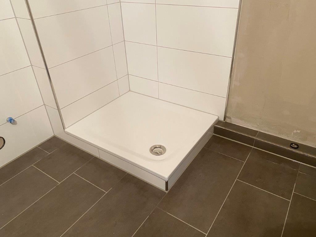 Zamontowany brodzik prysznicowy w narożniku łazienki, białe płytki ścienne i ciemna podłoga z płytek.