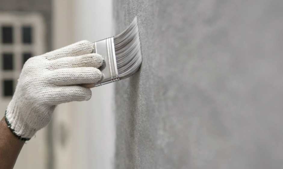 Dłoń w białej rękawicy malarskiej malująca szarą ścianę za pomocą pędzla.