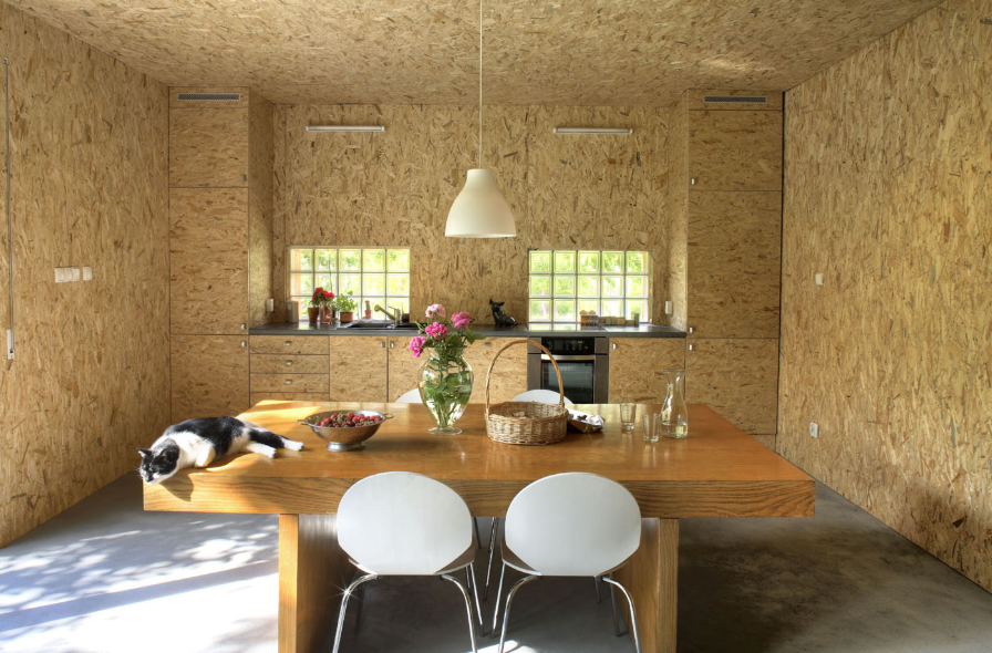 Wnętrze kuchni połączonej z jadalnią wykończonej płytami OSB, z drewnianym stołem, białymi krzesłami i centralną lampą wiszącą, doświetlonej naturalnym światłem z okien.