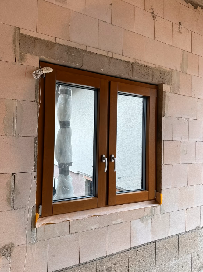 Dwuskrzydłowe okno z brązową ramą osadzone w ścianie z bloczków, z widocznymi klinami montażowymi oraz zabezpieczeniem ramy na etapie prac budowlanych.