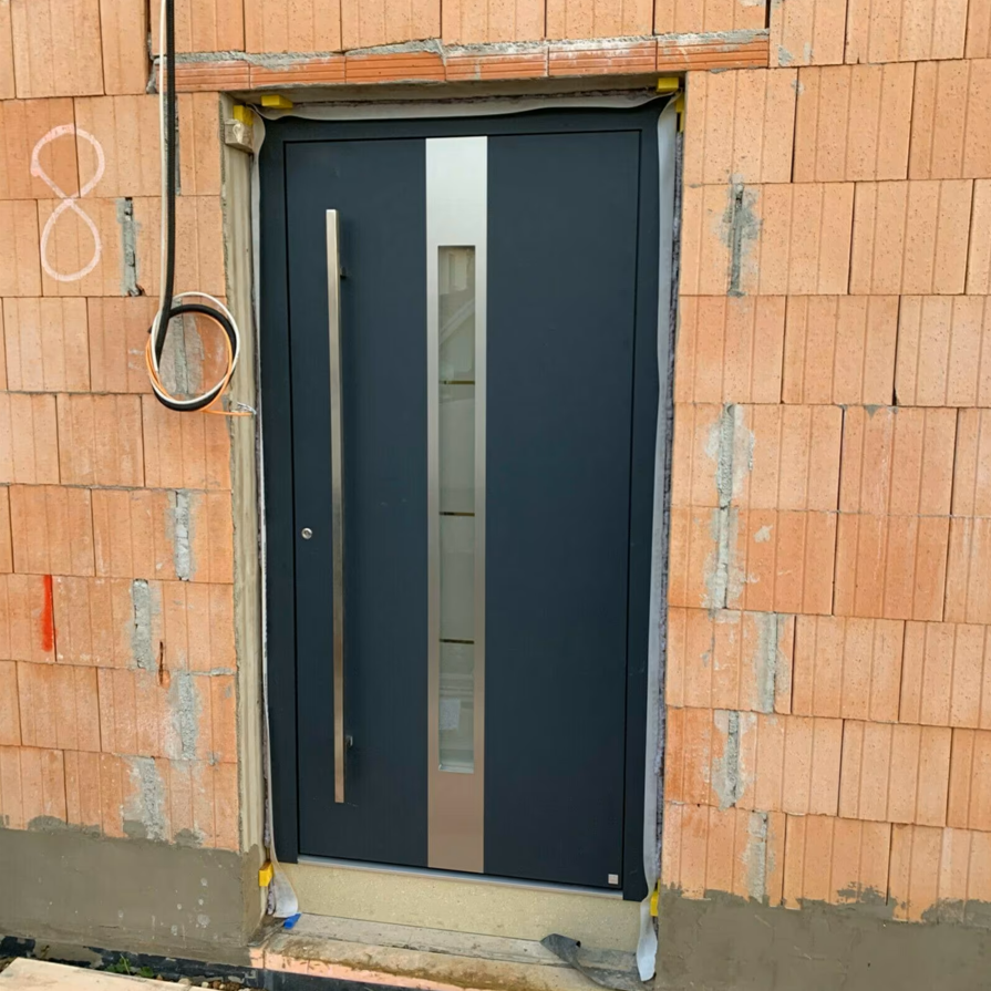 Drzwi zewnętrzne w kolorze antracytowym z pionowym przeszkleniem, zamontowane w otworze drzwiowym w ścianie z pustaków ceramicznych podczas prac budowlanych.