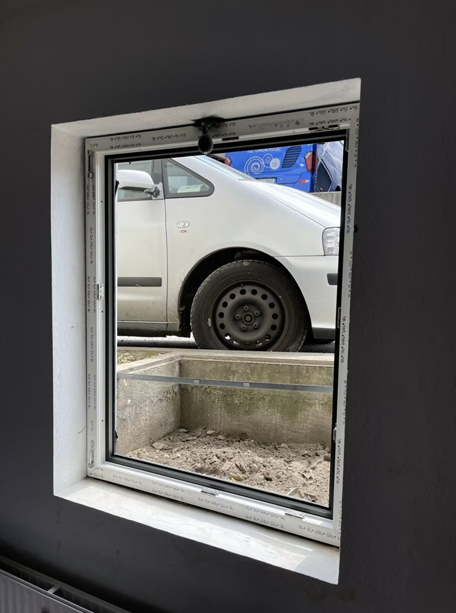 Okno piwniczne z białą ramą PVC osadzone w ścianie, widok od wewnątrz na studzienkę okienną przed wykończeniem wnęki.