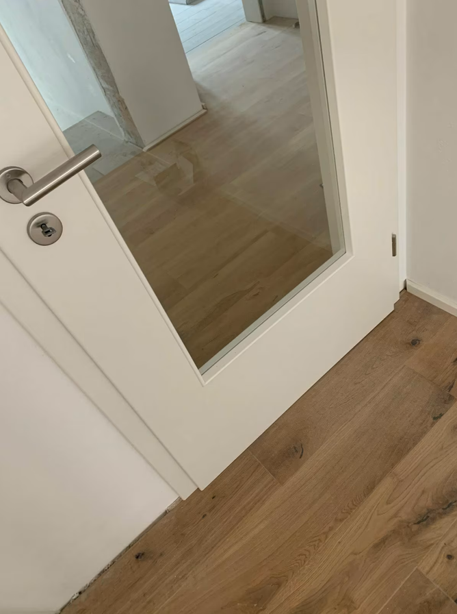 Fragment białych drzwi wewnętrznych z przeszkleniem oraz widok na drewnianą podłogę, prezentujący wykończenie detali po zakończeniu prac montażowych.