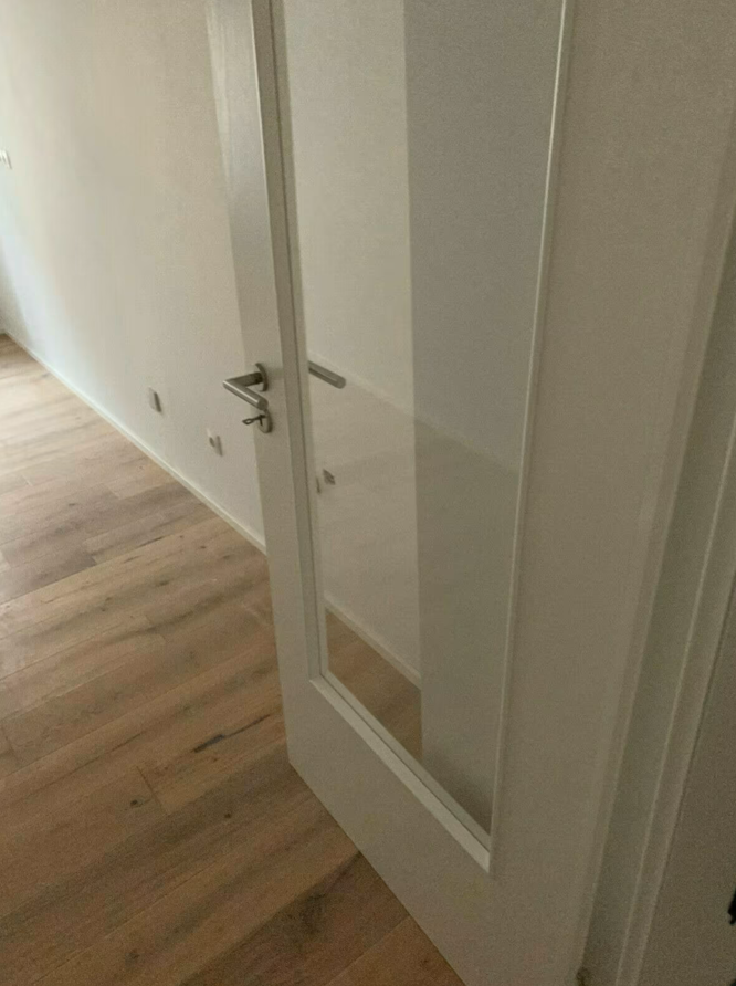 Białe skrzydło drzwiowe z dużym przeszkleniem i metalową klamką, osadzone w jasnym wnętrzu z drewnianą podłogą.