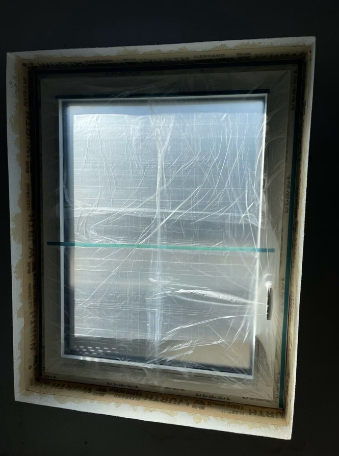 Osadzone okno z białą ramą PVC zabezpieczone folią ochronną, widok od wewnątrz przed wykonaniem obróbek wykończeniowych.