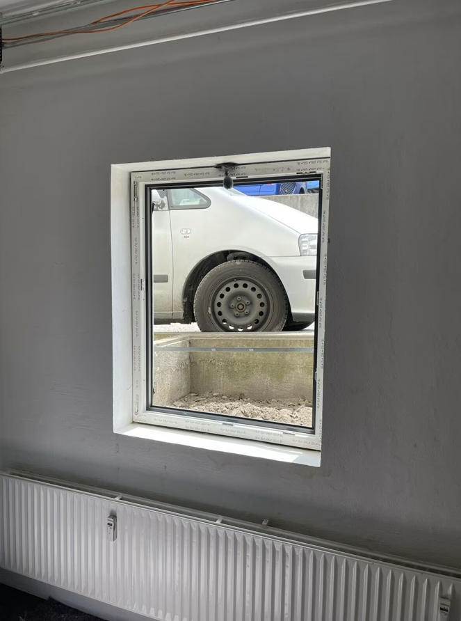 Widok od wewnątrz na zamontowane okno piwniczne z białą ramą PVC, osadzone w szarej ścianie, z widoczną wnęką zewnętrzną i zaparkowanym samochodem za oknem.