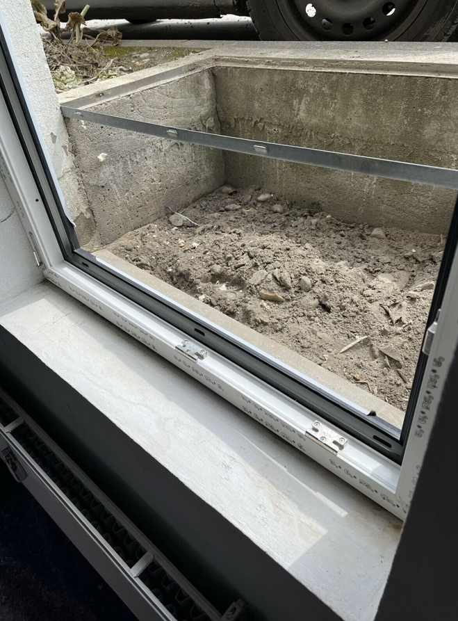 Widok od wewnątrz na okno piwniczne z białą ramą PVC, osadzone w ścianie budynku, z widoczną wnęką zewnętrzną, betonową obudową i nieutwardzonym gruntem przed oknem.