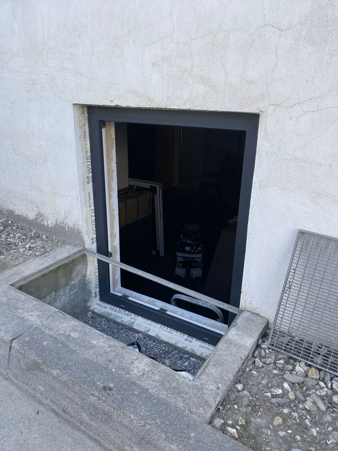 Okno piwniczne z ciemną ramą osadzone w ścianie budynku, widok z zewnątrz na etapie montażu, z widocznym otworem okiennym, ramą i prowizorycznym zabezpieczeniem.