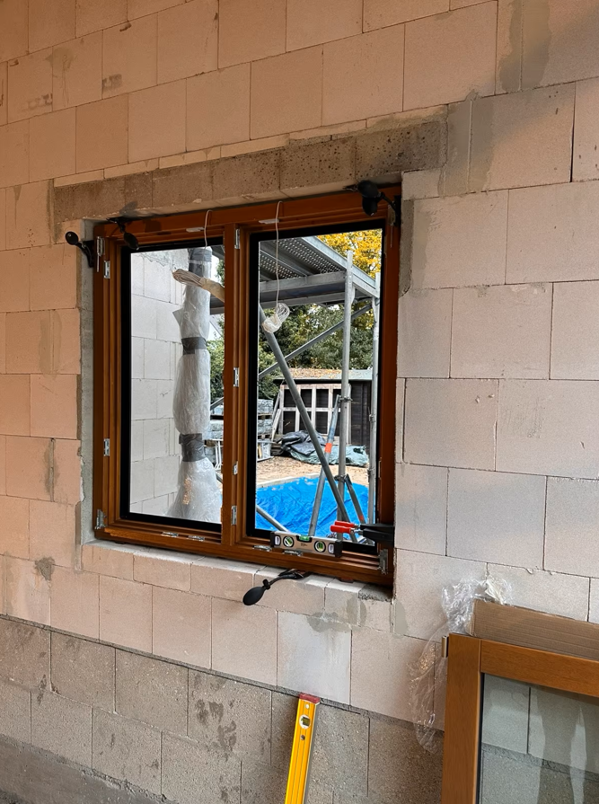 Dwuskrzydłowe okno z brązową ramą zamontowane w ścianie z bloczków, z widoczną poziomicą na dolnej ramie oraz elementami montażowymi i zabezpieczeniami podczas prac budowlanych.