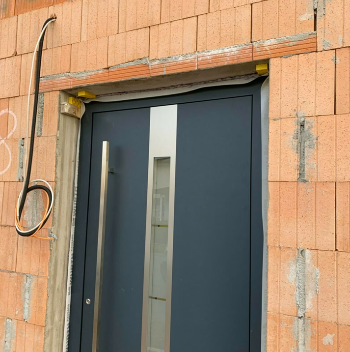 Nowoczesne drzwi zewnętrzne w kolorze antracytowym z pionowym przeszkleniem, osadzone w otworze drzwiowym w ścianie z pustaków ceramicznych podczas montażu.